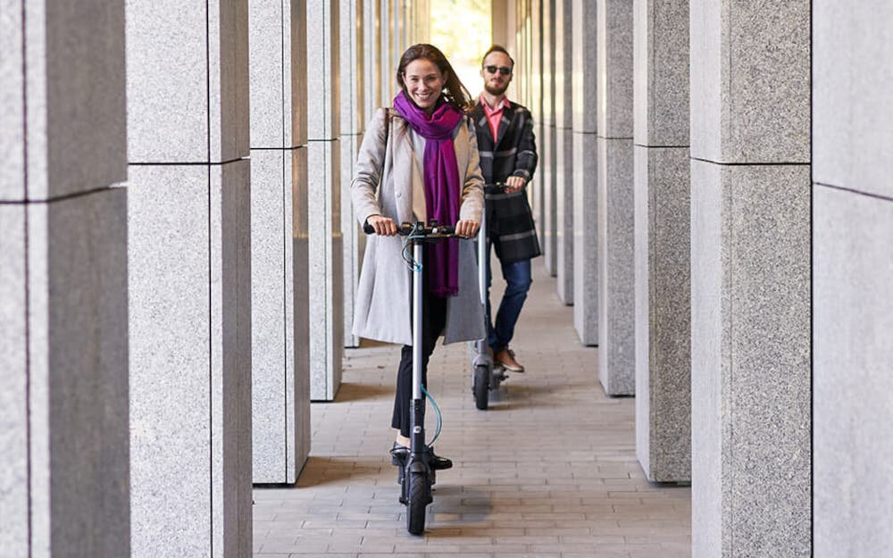 Hulajnoga elektryczna MOTUS Scooty 10 Lite Gen 4 30km 350W Amortyzacja 10 cali Szary dwie osoby jadą na hulajnodze jedna za drugą chodnik budynek 3 tryby jazdy tryb pieszy prędkość do 6 km/h tryb standardowy prędkość do 15 km/h tryb sportowy prędkość do 20 km/h