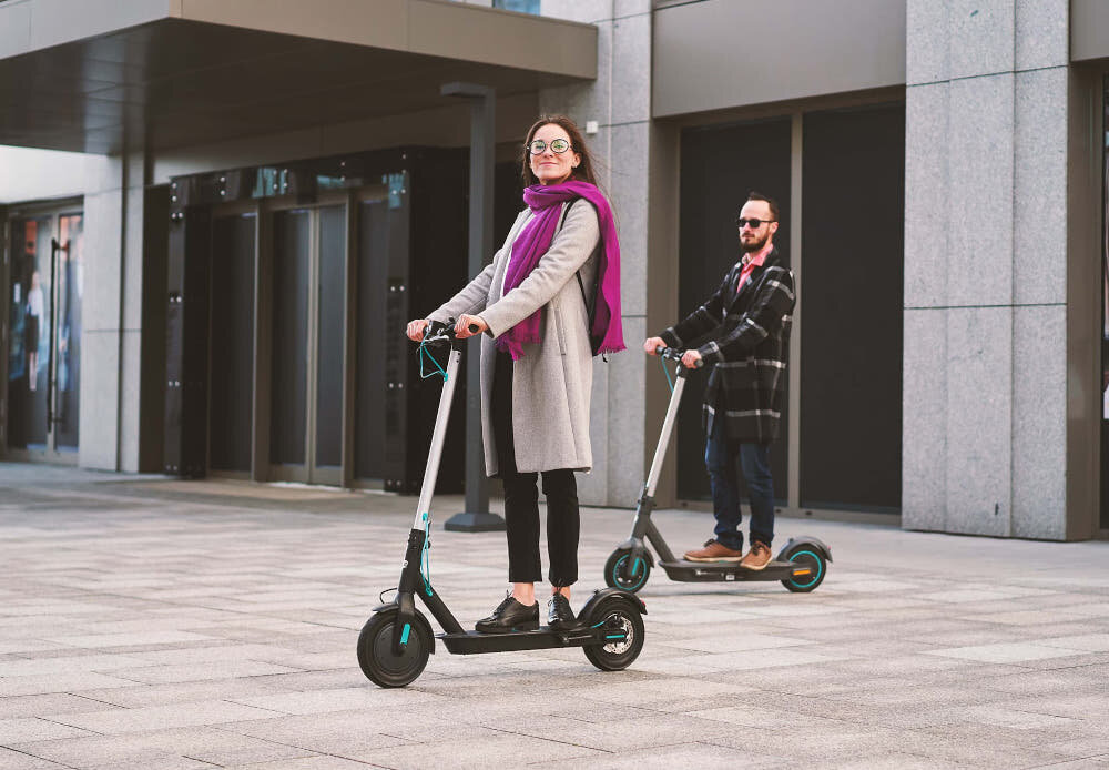 Hulajnoga elektryczna MOTUS Scooty 10 Lite Gen 4 30km 350W Amortyzacja 10 cali Szary dwie osoby jadą na hulajnogach chodnik w tle budynek certyfikat IP54 bezpieczne podróżowanie w różnych warunkach atmosferycznych odporność na kurz, brud i zachlapania nie można jej zanurzać bezpośrednio w wodzie