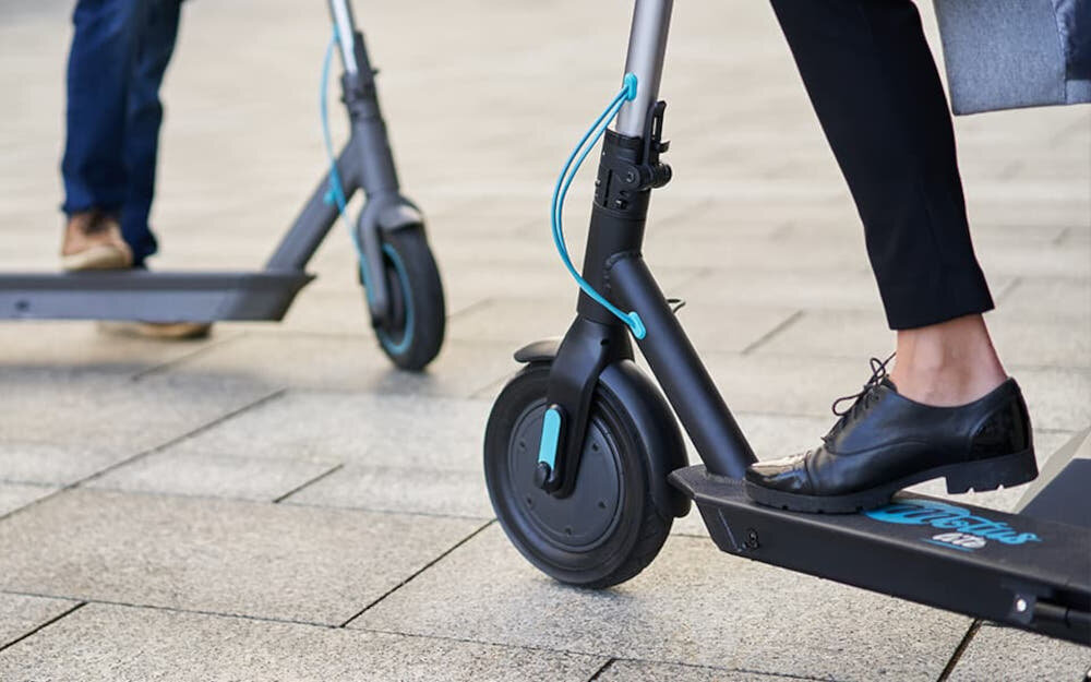 Hulajnoga elektryczna MOTUS Scooty 10 Lite Gen 4 30km 350W Amortyzacja 10 cali Szary zbliżenie na przednie koło osoba opiera stopę o deck chodnik 10-calowe koła dętkowe o niskim oporze toczenia duża przyczepność płynna i oszczędna jazda absorbują wstrząsy i nierówności terenu stabilna i wygodna jazda duża elastyczność podczas jazdy