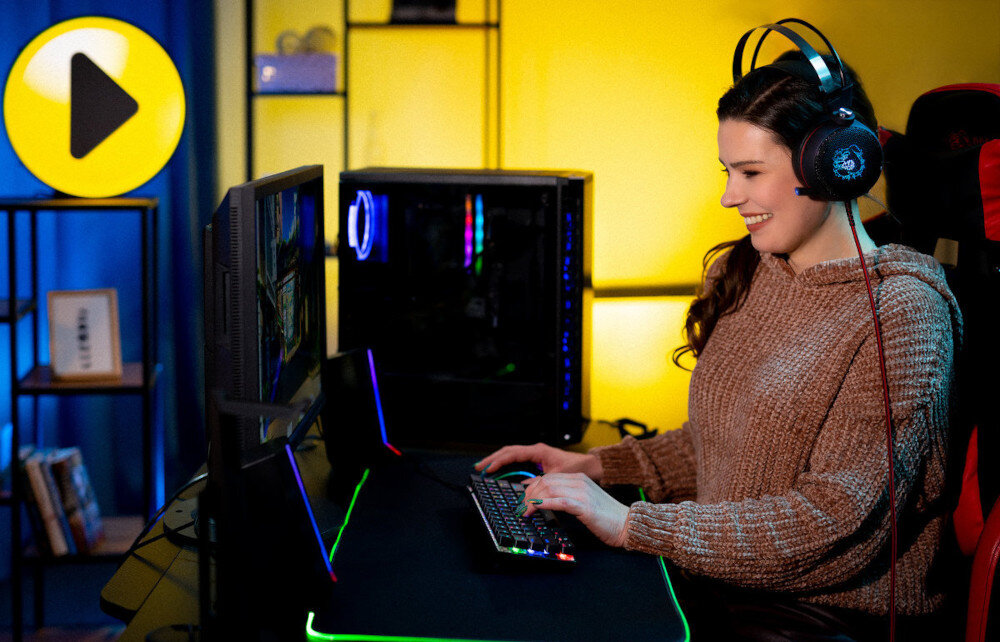 Komputer MAD DOG GeForce RTX5070 DLSS 4 Reflex Edition 2 ENDORFY500ARGB-A33DR32 Widok od boku, kobieta w słuchawkach gamingowych siedząca przy biurku grająca w grę, w tle komputer wygląd opis