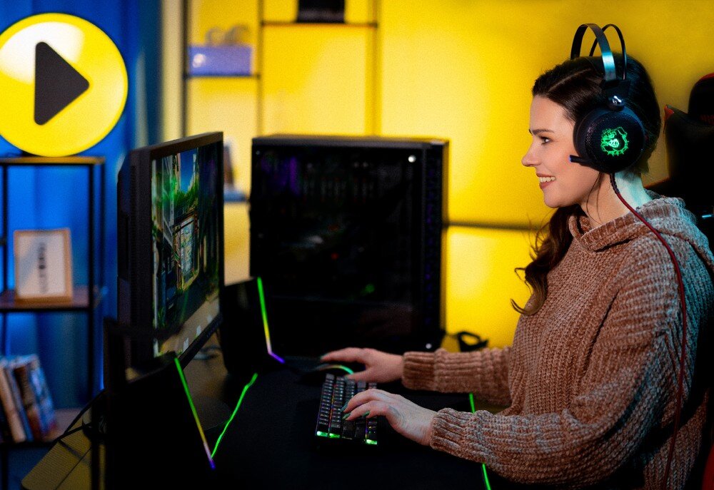 Komputer MAD DOG Fortnite Ready FR-01DB32V2 Kobieta w słuchawkach grająca na komputerze stacjonarnym. Siedzi przy biurku z podświetlaną klawiaturą i monitorem, w tle niebiesko-żółte oświetlenie i regał, opis