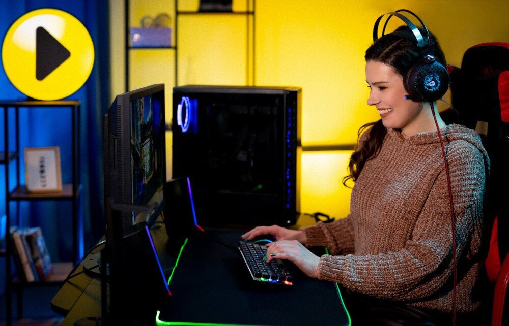 Komputer MAD DOG Fortnite Ready FR-03WR32WHV2 Kobieta w słuchawkach siedzi przy biurku i gra na komputerze. Monitor oraz klawiatura mają podświetlenie RGB. W tle obudowa komputera widoczna obok monitora z podświetlonym wnętrzem, opis