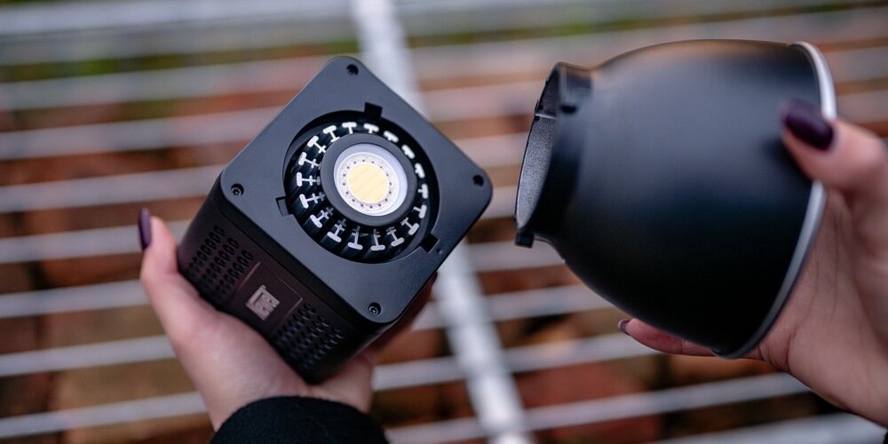 Kobiece dłonie na tle metalowej kratki zakładają na lampę LED Newell Tara 40 RGB z systemem Bowens czaszę.