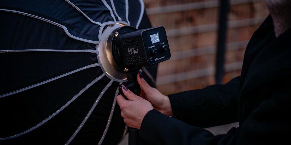 Kobiece dłonie przymocowują softbox do lampy Tara marki Newell na tle ceglastego budynku.