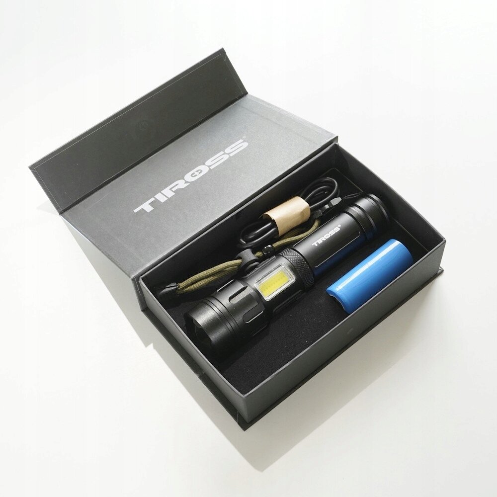 Czarna latarka LED z napisem \'TIROSS\' znajduje się w otwartym pudełku. Obok latarki leży niebieski akumulator i czarny kabel USB.