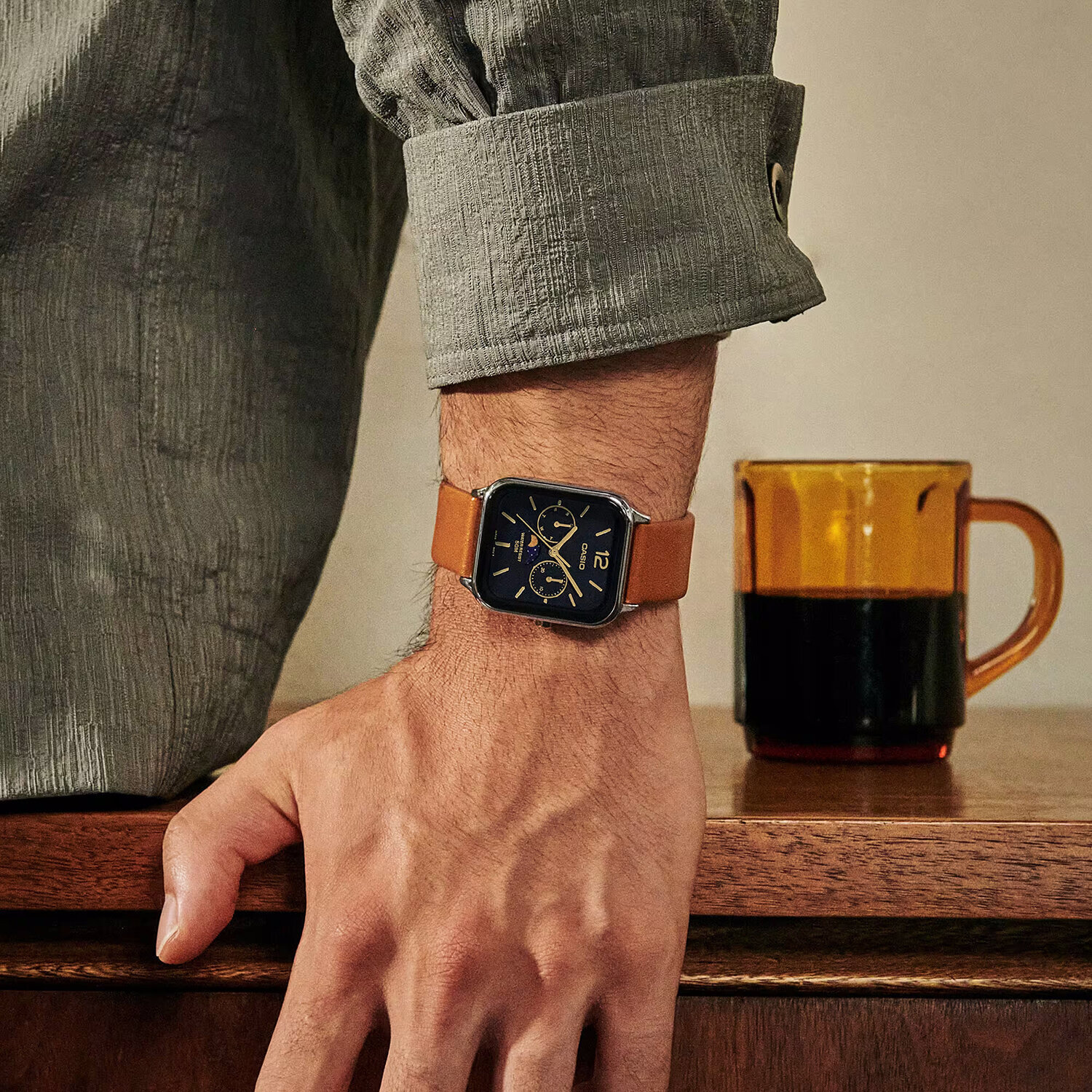 Osoba nosi zegarek z brązowym paskiem i ciemną tarczą z widocznym logo Casio. Obok na drewnianym blacie stoi szklany kubek z ciemnym napojem.