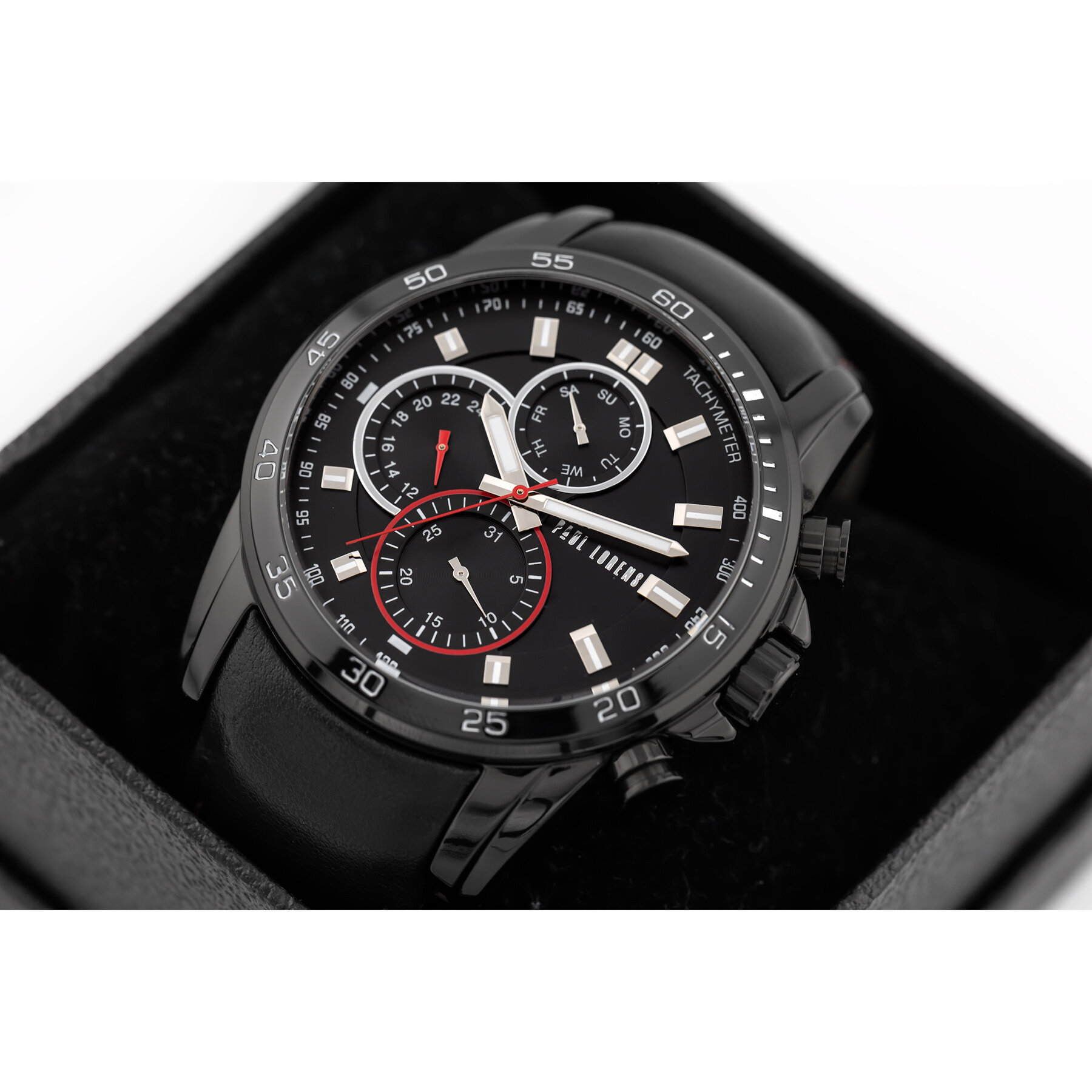 Czarny zegarek z trzema mniejszymi tarczami i białymi indeksami, umieszczony w pudełku. Na tarczy widoczny jest napis \'TACHYMETER\'.