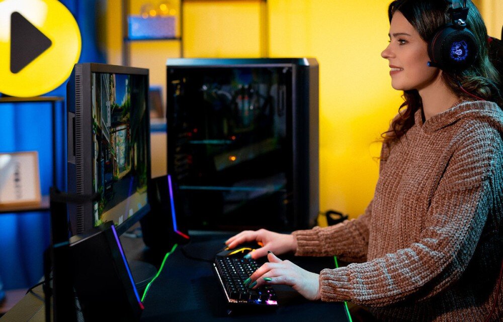 Komputer MAD DOG GeForce RTX5060Ti DLSS 4 Reflex Edition 2 BQ501LX-A01WR64WH Młoda kobieta w słuchawkach na uszach siedząca z profilu przy biurku, obsługująca podświetlaną klawiaturę i mysz, z widocznym monitorem i komputerem w tle