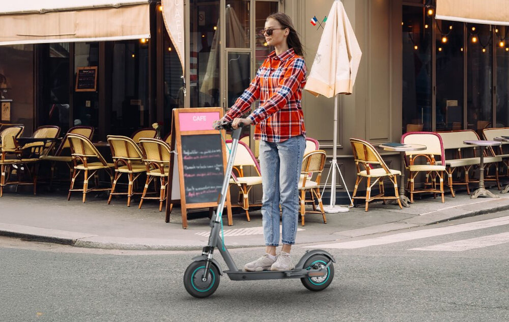 Hulajnoga elektryczna MOTUS Scooty 10 Lite Gen 6 30km 350W Amortyzacja 10 cali Szary miasto kobieta jedzie na hulajnodze w tle restauracja 3 tryby jazdy przycisk POWER umieszczonym na wyświetlaczu tryb pieszy prędkość do 6 km/h tryb standardowy prędkość do 15 km/h tryb sportowy prędkość do 20 km/h