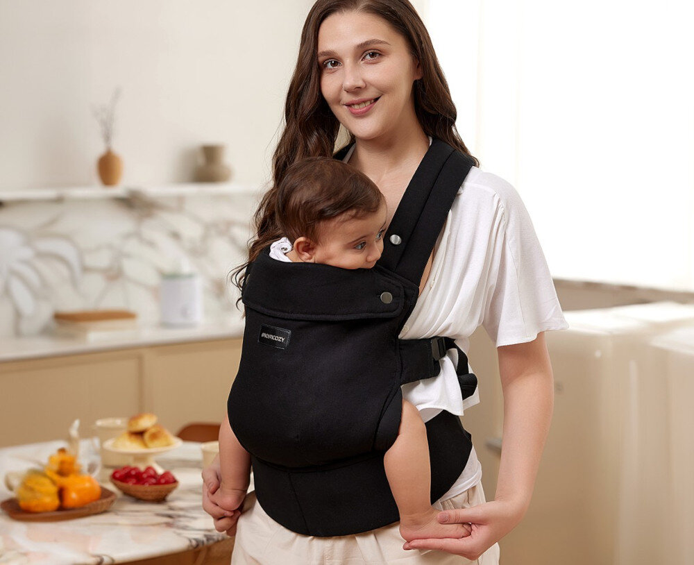 Osoba dorosła nosi dziecko w czarnym nosidełku z widocznym logo \'MOMCOZY\'. W tle znajduje się stół z owocami i pieczywem oraz jasna kuchnia.