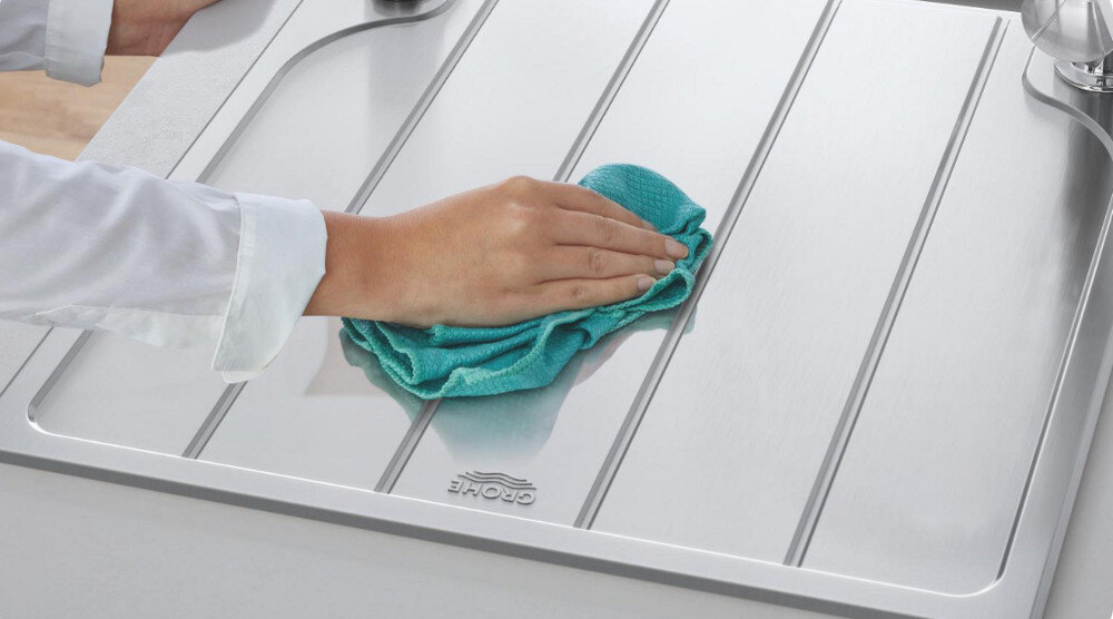 Zlewozmywak jednokomorowy z ociekaczem GROHE K200 31565SD1 Stal szlachetna 50x86 + bateria stojąca Eurosmart 3328100311 Na zdjęciu widoczna jest dłoń w białym rękawie, przecierająca turkusową ściereczką stalowy ociekacz zlewozmywaka. Gładka powierzchnia ze szczotkowanej stali nierdzewnej błyszczy i podkreśla elegancki wygląd oraz łatwość czyszczenia gładka powierzchnia stal szlachetna łatwe czyszczenie higiena właściwości antybakteryjne kontakt z żywnością