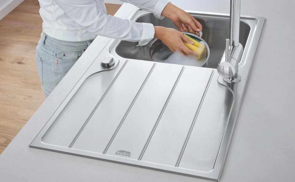 Zlewozmywak jednokomorowy z ociekaczem GROHE K500 31571SD1 Stal szlachetna Na zdjęciu widać stalowy zlewozmywak jednokomorowy z ociekaczem, osadzony w blacie o ciemnoszarej powierzchni. W centralnej części ociekacza znajduje się chromowana, jednouchwytowa bateria stojąca, a w tle widoczne są minimalistyczne akcesoria kuchenne na białej półce.