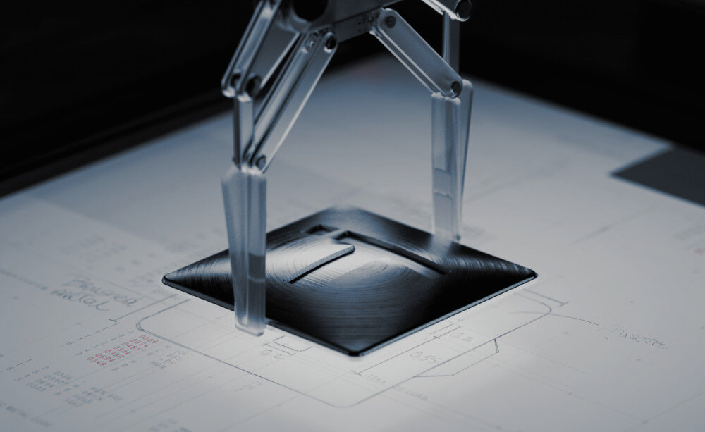 Piekarnik TEKA NEO HSB 6350 SS Zdjęcie pokazuje precyzyjne ramię pomiarowe lub testowe, które dotyka metalowego elementu o stożkowym kształcie, umieszczonego na powierzchni z rysunkami technicznymi. Oświetlenie punktowe wydobywa strukturę metalu i sugeruje proces kontroli jakości lub testowania komponentu logo T symbol znak elegancja znak rozpoznawczy firmy jakość detal styl kuchnia