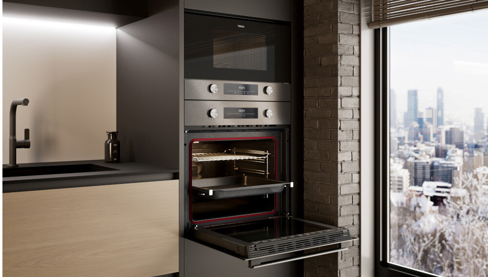 Piekarnik TEKA NEO HSB 6350 SS Zzdjęcie przedstawia nowoczesną kuchnię z piekarnikiem TEKA w zabudowie, którego drzwi są otwarte, a blacha delikatnie wysunięta na prowadnicach. Przestrzeń utrzymana jest w połączeniu ciemnych frontów, jasnego blatu oraz dużego okna z widokiem na panoramę miasta 70 litrów przestrzeni to gwarancja swobody podczas pieczenia