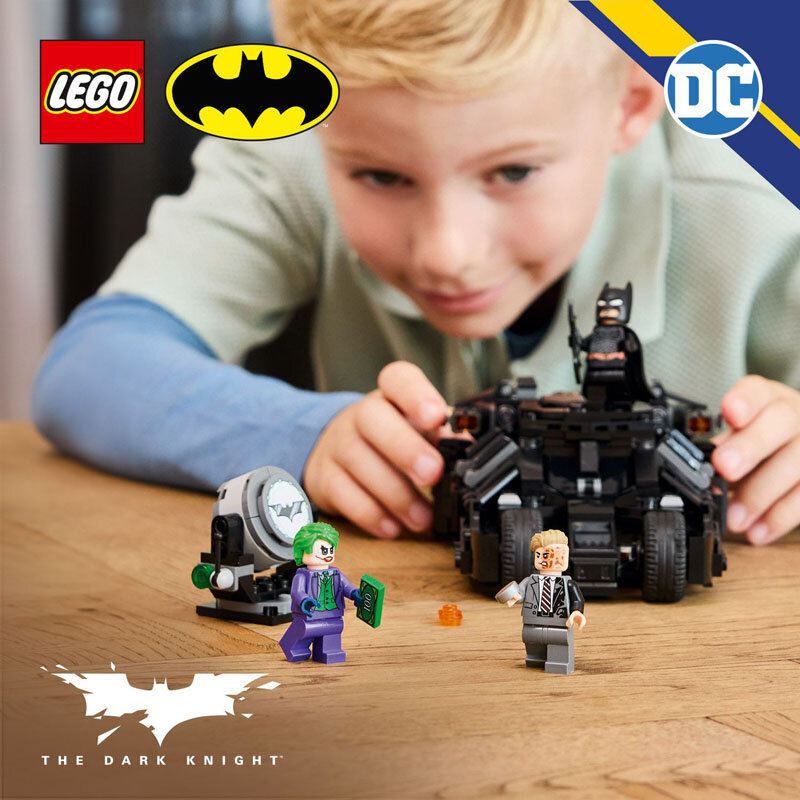 Na drewnianym stole znajdują się figurki LEGO przedstawiające postacie z uniwersum Batmana oraz pojazd Batmana. W tle widoczne są logotypy LEGO, Batman i DC, a także napis \'The Dark Knight\'.