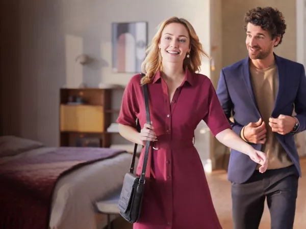 Dwie osoby znajdują się w jasnym pomieszczeniu, jedna z nich trzyma torbę na ramieniu. W tle widoczny jest łóżko i półka z książkami.