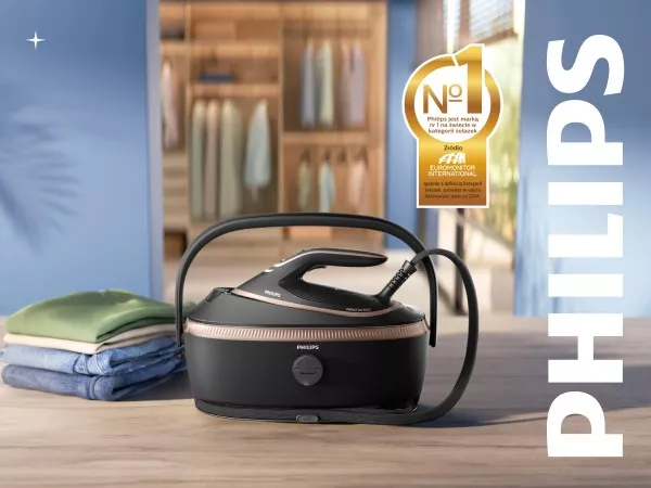 Na drewnianym stole znajduje się generator pary marki PHILIPS z czarną podstawą i uchwytem. Obok widoczny jest stos złożonych ubrań, a w tle znajduje się szafa z wiszącymi ubraniami.