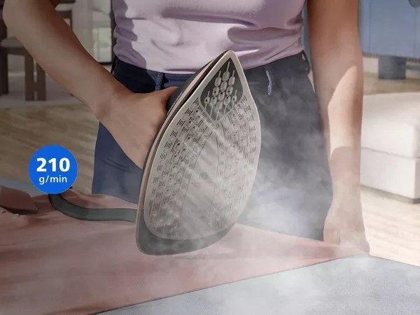 Osoba prasuje tkaninę za pomocą żelazka, z którego wydobywa się para. Na obrazie widoczny jest napis \'210 g/min\'.