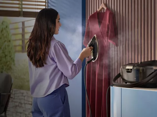 Osoba używa generatora pary do prasowania wiszącej sukienki. W tle widoczne są elementy wnętrza, takie jak ściana i okno.