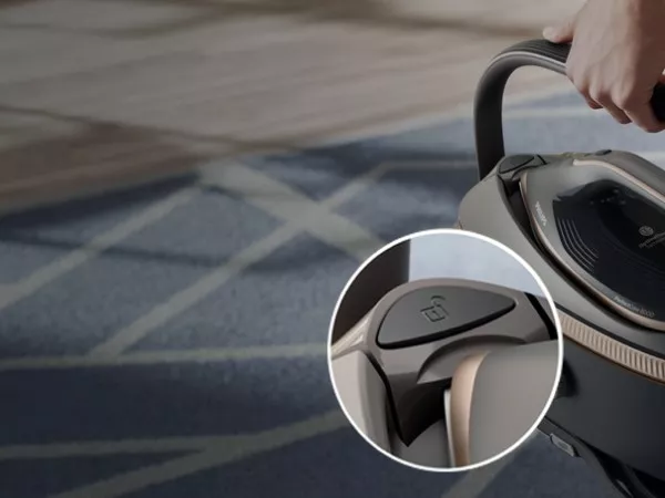 Zdjęcie przedstawia rękę trzymającą uchwyt urządzenia, prawdopodobnie generatora pary, na tle dywanu z geometrycznym wzorem. W okrągłym zbliżeniu widoczny jest przycisk z symbolem kłódki.