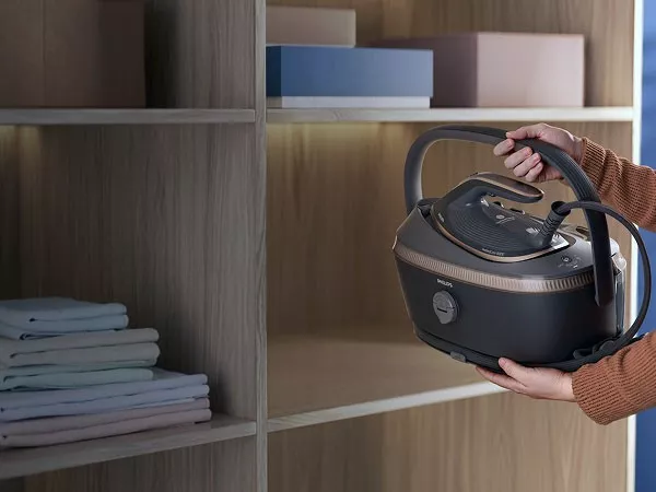 Osoba trzyma generator pary marki Philips w pobliżu półki z ułożonymi ubraniami. Urządzenie ma ciemną obudowę z jasnymi akcentami i widoczne logo Philips.