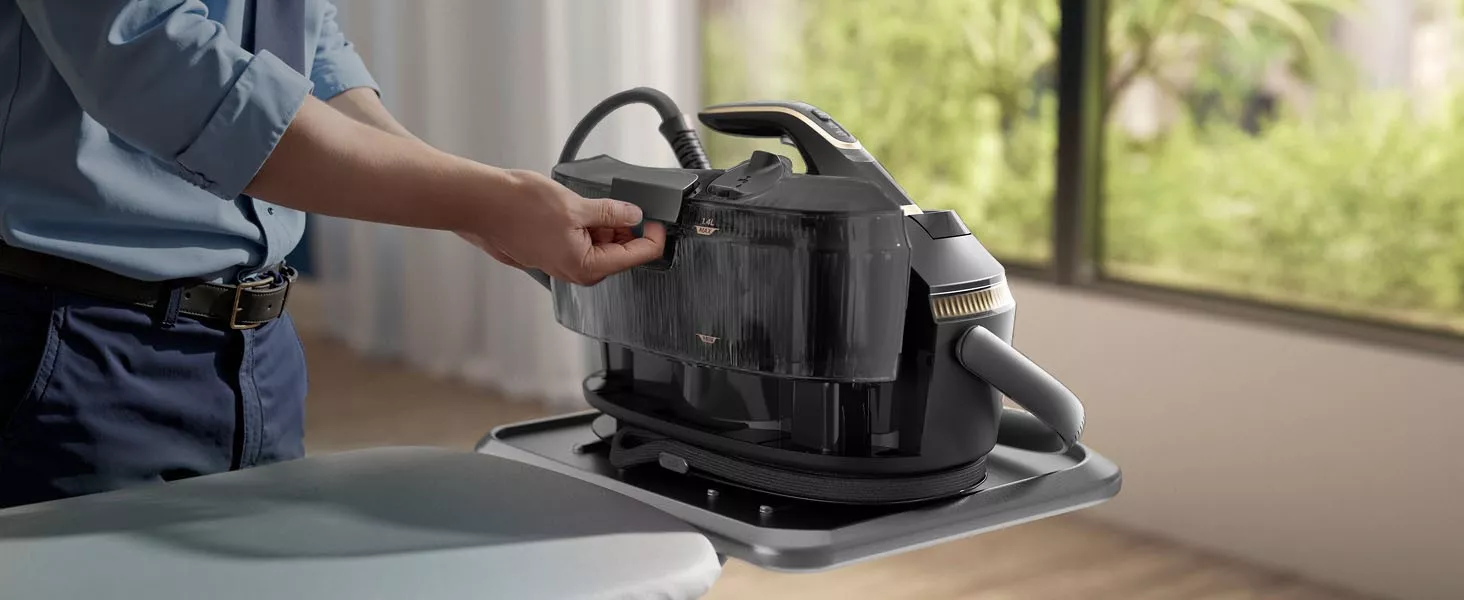 Osoba trzyma generator pary umieszczony na desce do prasowania. W tle widoczne jest rozmyte okno z zielenią na zewnątrz.