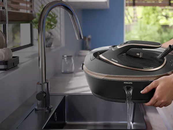 Osoba trzyma generator pary PHILIPS nad zlewem, z którego wylewa się woda. W tle widoczne są elementy kuchni, takie jak kran i okno.