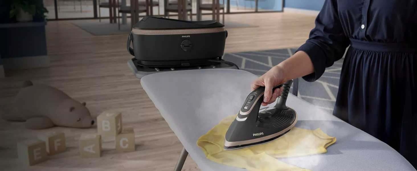 Osoba prasuje żółtą tkaninę za pomocą generatora pary marki PHILIPS na desce do prasowania. W tle widać pluszowego misia i klocki z literami E, F, A, B, C.