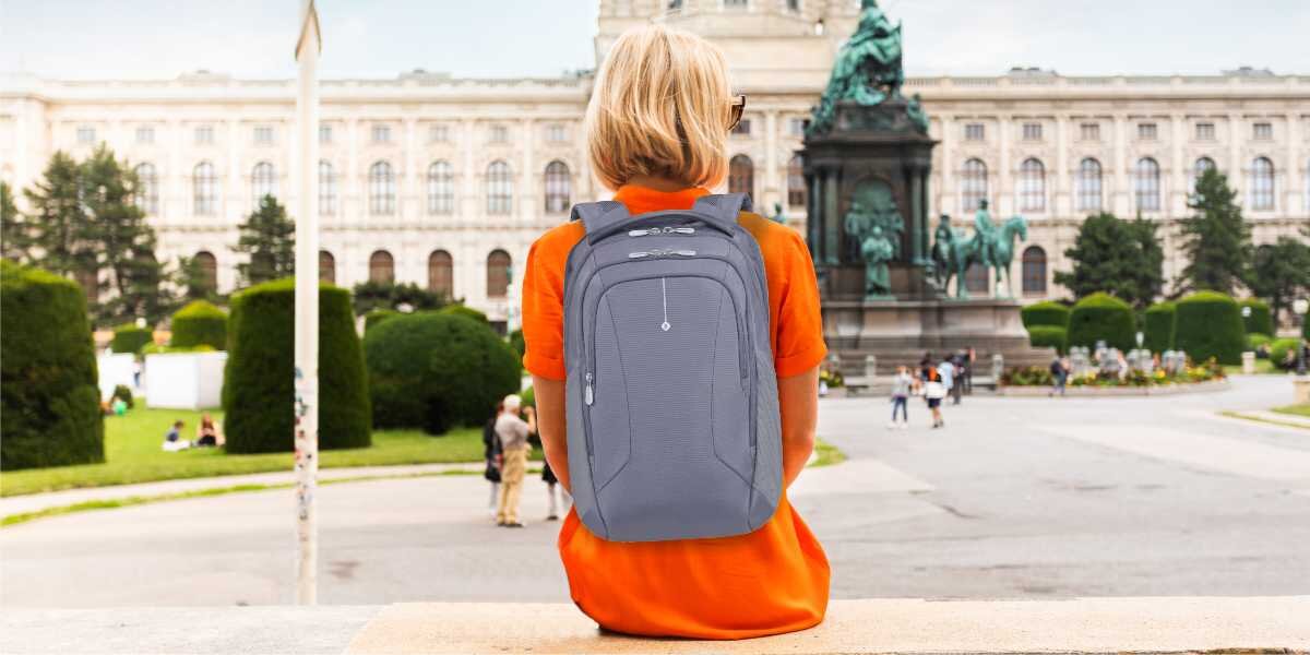 Plecak na laptopa SAMSONITE Guardit Classy 2.0 15.6 cali Niebieski Kobieta w pomarańczowej sukience z plecakiem Samsonite na plecach siedzi na murku w Wiedniu na tle zabytkowego budynku i pomnika.