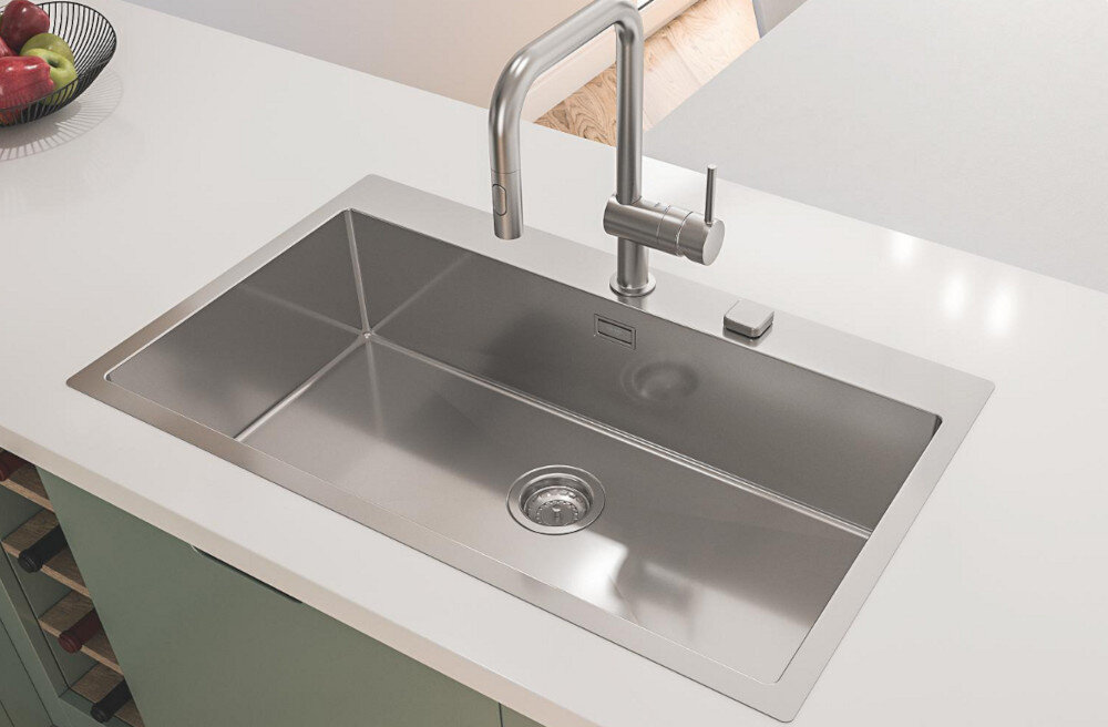 Zlewozmywak jednokomorowy GROHE K800 31584SD1 Stal szlachetna Na zdjęciu widać stalowy zlewozmywak jednokomorowy z ociekaczem, osadzony w blacie o ciemnoszarej powierzchni. W centralnej części ociekacza znajduje się chromowana, jednouchwytowa bateria stojąca, a w tle widoczne są minimalistyczne akcesoria kuchenne na białej półce.