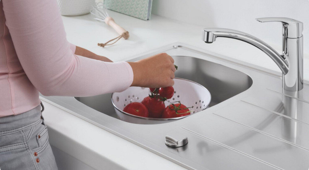 Zlewozmywak jednokomorowy GROHE K800 31584SD1 Stal szlachetna Na zdjęciu osoba w różowej bluzce płucze czerwone pomidory na gałązce w białym durszlaku umieszczonym w komorze stalowego zlewozmywaka. Obok widoczny jest ociekacz ze zintegrowanym pokrętłem oraz chromowana bateria kuchenna na tle jasnego blatu i akcesoriów kuchennych zlewozmywak jednokomorowy duża komora ociekacz ergonomia wygoda użytkowania