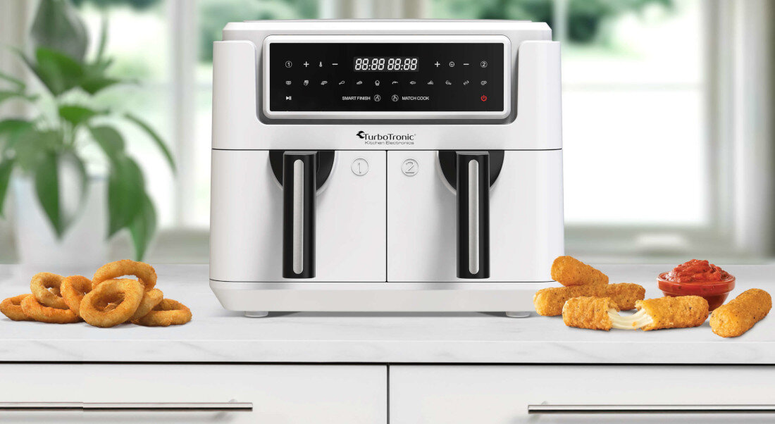 Na blacie kuchennym stoi biała frytkownica beztłuszczowa z podwójnym koszem marki TurboTronic. Obok urządzenia znajdują się krążki cebulowe i paluszki serowe z sosem.