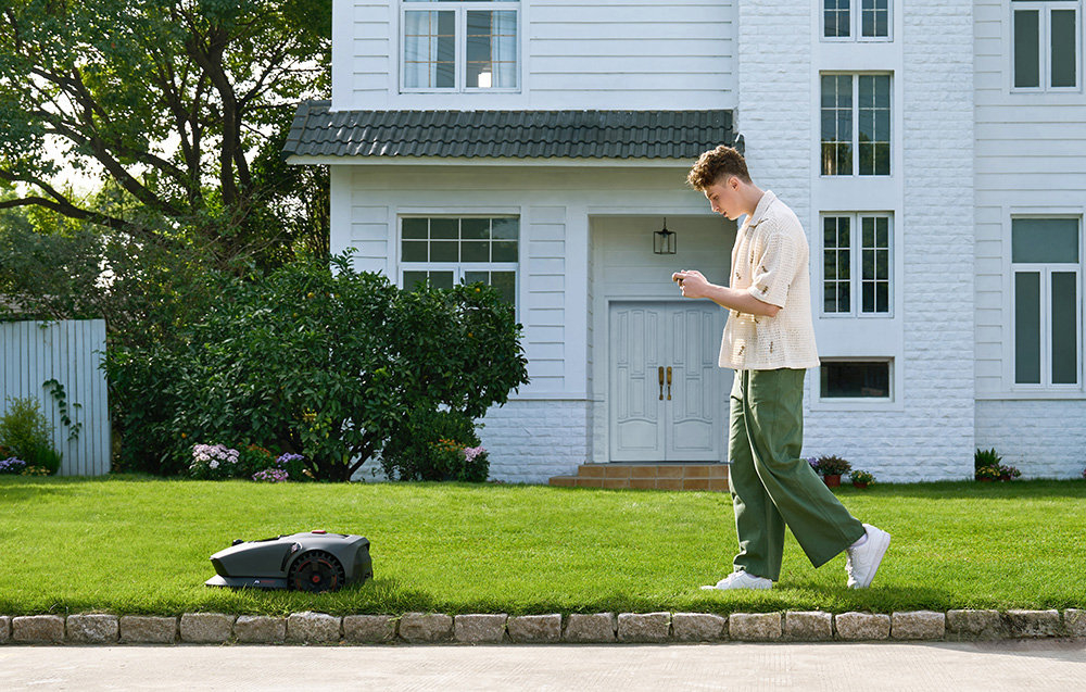 Robot koszący DREAME Mova 1000 robot na trawie za nim osoba z telefonem w dłoni w tle dom i rośliny wyznacza wirtualne granice aplikacja MOVAHome wielostrefowe mapy harmonogramy pracy wyłączać obszary kontrolę koszenia bezpośrednio z poziomu telefonu bezpieczeństwo Twoich kwiatów wbudowany moduł Wi-Fi