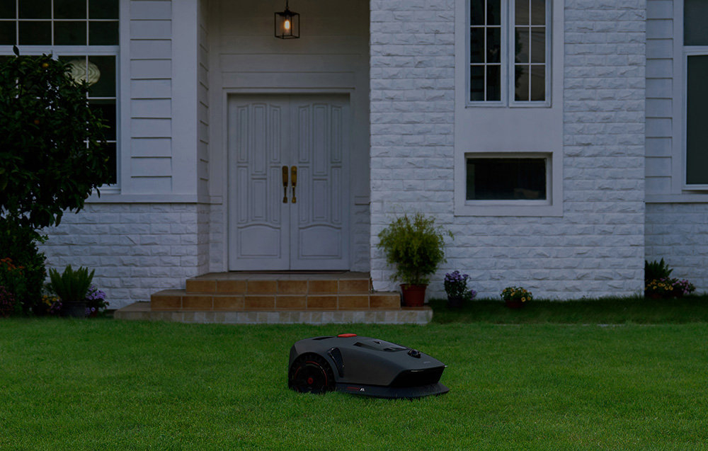 Robot koszący DREAME Mova 1000 robot na trawie w tle biały dom wieczorna pora poziom hałasu nie przekracza 60 dB akumulator ekologiczne rozwiązanie akumulator o pojemności 2,5 Ah i napięciu 18 V czas pracy około 50 minut na jednym ładowaniu
