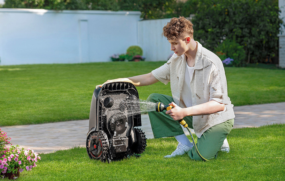 Robot koszący DREAME Mova 1000 osoba myje wężem ogrodowym  robota w tle trawnik rośliny i płot wodoodporność IPX6 pracuje w każdych warunkach atmosferycznych łatwy w czyszczeniu automatycznie wraca do stacji dokującej do 90% w zaledwie 40 minut pełne ładowanie w około 110 minut