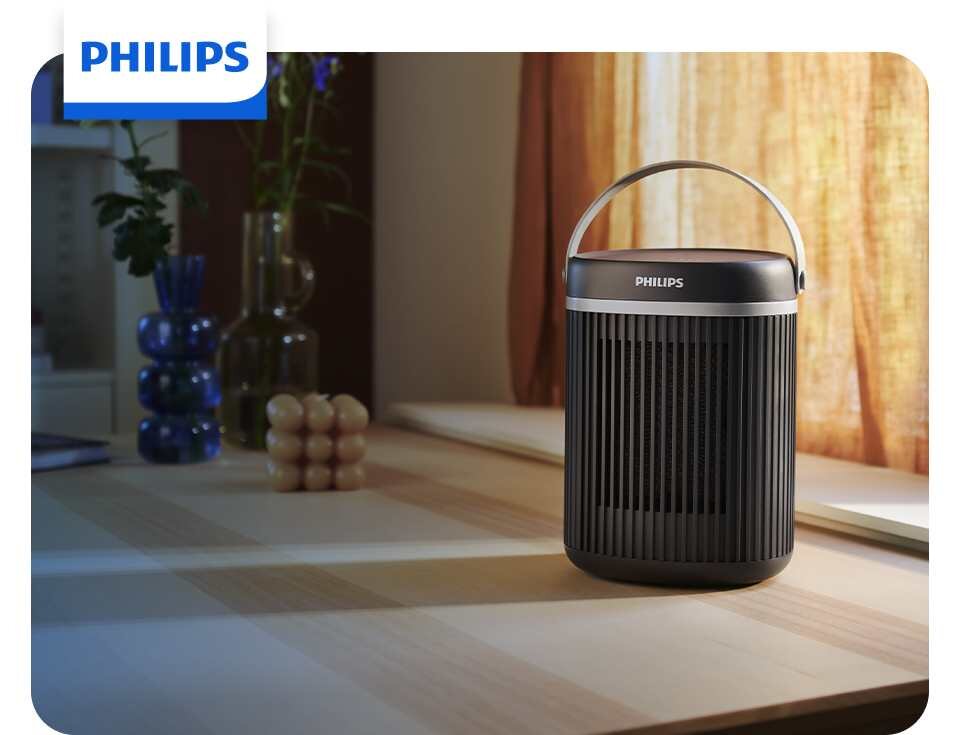 Na drewnianym stole stoi czarny grzejnik ceramiczny z logo PHILIPS na górze. W tle widoczne są zasłony i dekoracyjne wazony.