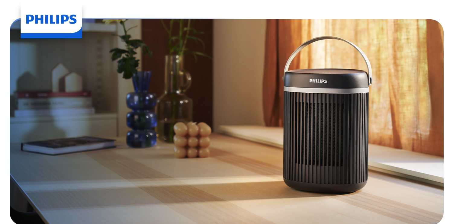 Na drewnianym stole stoi czarny grzejnik ceramiczny z logo PHILIPS, z uchwytem na górze. W tle widoczne są zasłony, książki i dekoracyjne wazony.