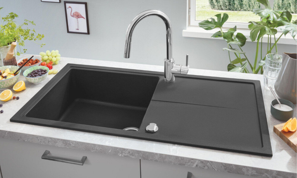 Zlewozmywak jednokomorowy z ociekaczem GROHE K400 31641AP0 Czarny jednokomorowy zlewozmywak wpuszczany w blat z wysoką, chromowaną baterią kuchenną. Wokół stoją produkty spożywcze i dodatki kuchenne tworzące estetyczną aranżację