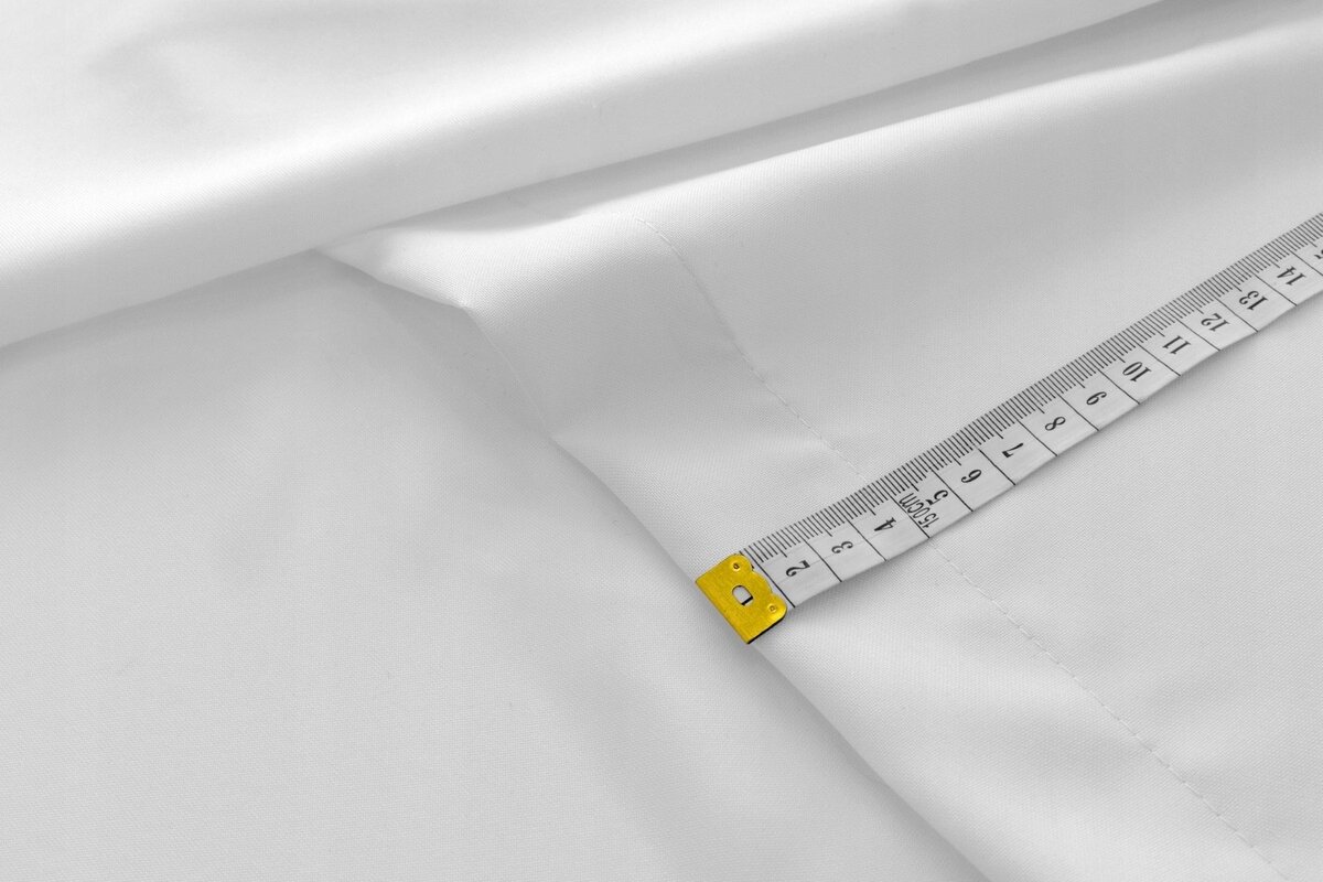 Biały materiał złożony na stole, obok którego leży miarka krawiecka z widocznymi centymetrami. Miarka ma żółty uchwyt i pokazuje długość do 15 cm.