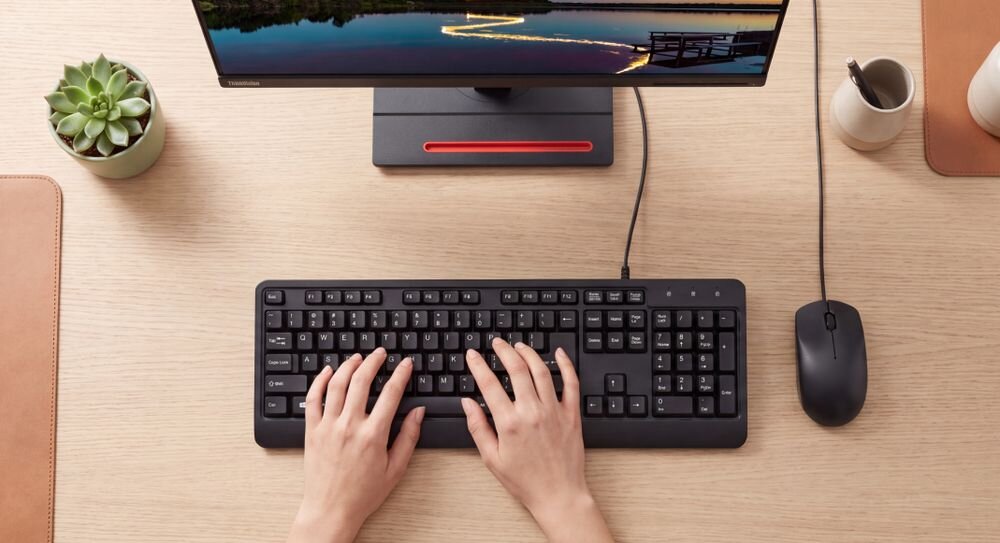 LENOVO 160 Wired Combo Trwałość klawiatury do 8 milionów naciśnięć klawiszy przyciski myszy 3 milionów lewego i prawego przycisku - Widok z góry na dłonie piszące na czarnej klawiaturze na zorganizowanym drewnianym biurku z monitorem