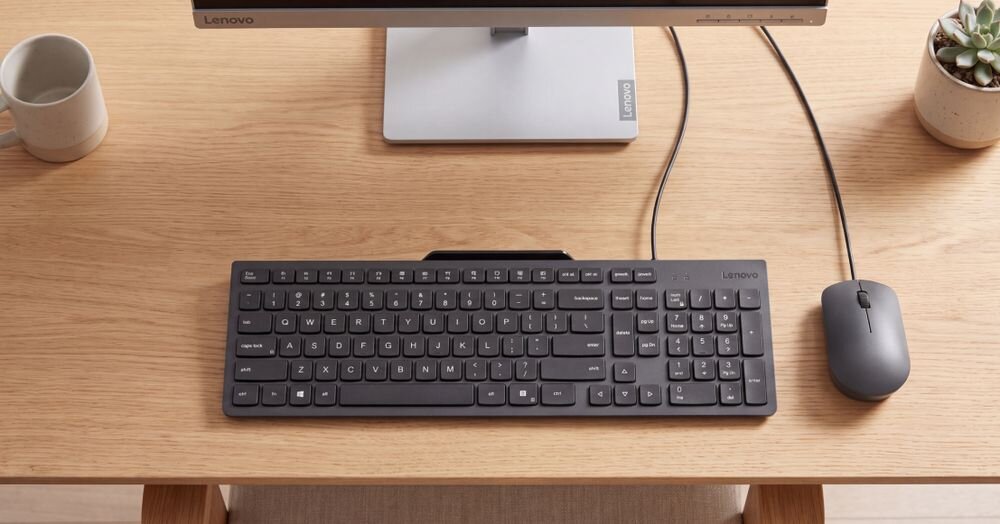 Na drewnianym biurku znajduje się czarna klawiatura i myszka przewodowa obok monitora. Na klawiaturze widoczne jest logo \'Lenovo\'.