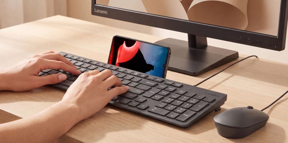 Na biurku znajduje się klawiatura i myszka przewodowa obok monitora z logo Lenovo. Osoba pisze na klawiaturze, a przed nią stoi telefon z wyświetlonym obrazem.