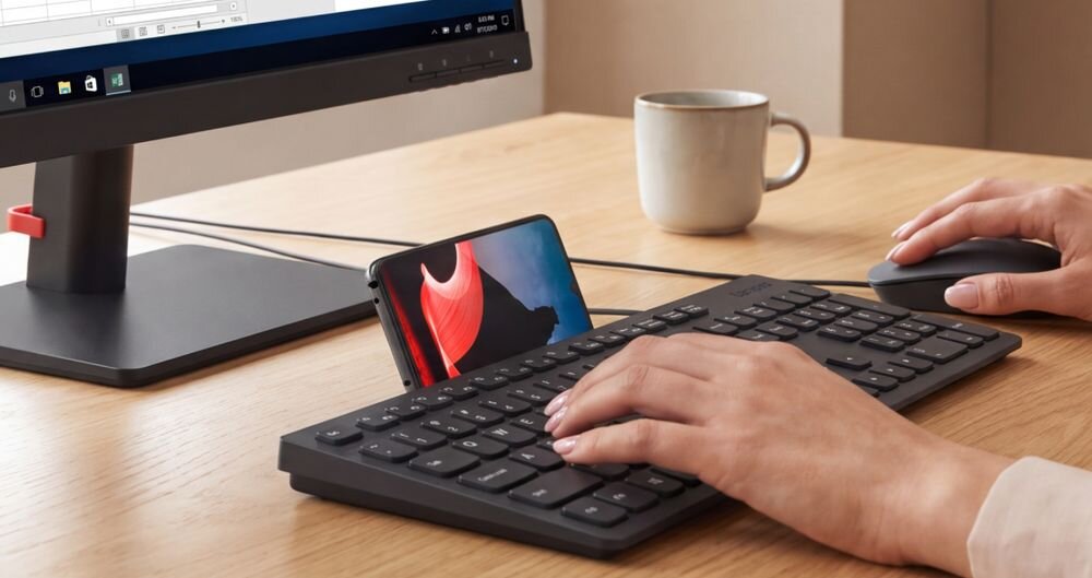 Na drewnianym biurku znajduje się monitor, klawiatura, myszka oraz telefon w uchwycie. Osoba pisze na klawiaturze, a w tle stoi kubek.