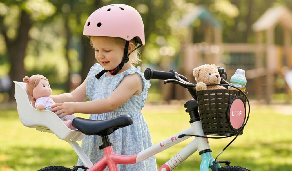 Dziecko w kasku bawi się lalką na rowerze dziecięcym w kolorach biało-różowo-miętowym. Na koszyku roweru widoczne jest logo \'LOOFI\'.