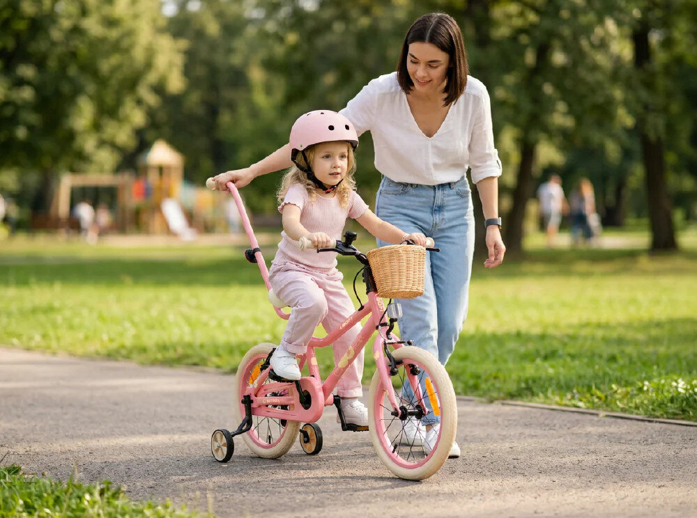 Dziewczynka w różowym kasku jedzie na różowym rowerze dziecięcym z koszykiem, obok niej idzie kobieta. W tle widać zielony park.