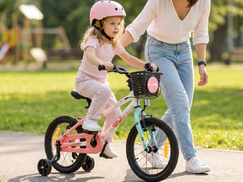Dziecko w różowym kasku jedzie na biało-różowo-miętowym rowerze dziecięcym z bocznymi kółkami, obok stoi osoba dorosła. W tle widać park z zielenią.