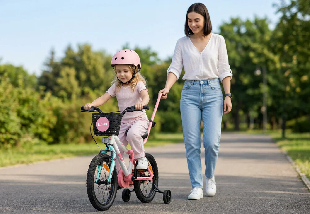 Dziewczynka w różowym kasku jedzie na biało-różowo-miętowym rowerze dziecięcym z bocznymi kółkami, a obok idzie kobieta trzymająca drążek przymocowany do roweru. W tle widać zielone drzewa i ścieżkę w parku.