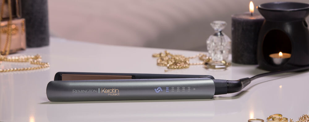 Na białym stole leży prostownica z napisem \'Remington Keratin Protect\'. W tle widoczne są świece, biżuteria i dekoracyjne przedmioty.