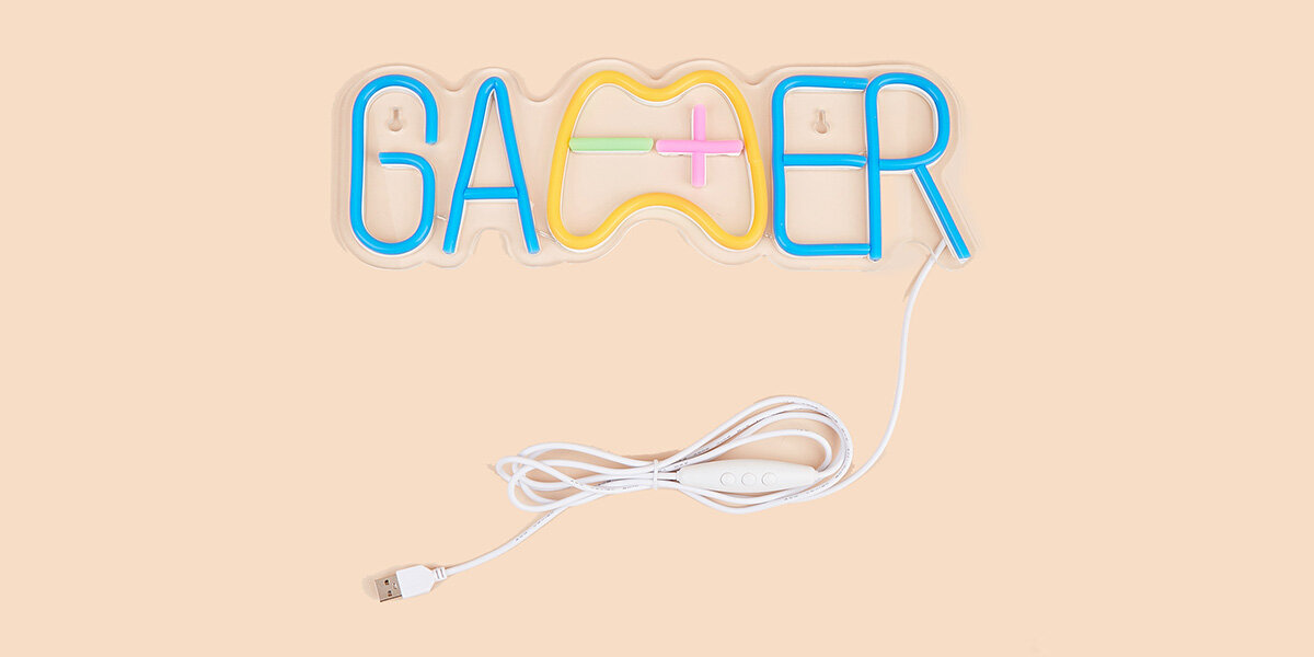 Neonowy napis \'GAMER\' w kolorach niebieskim, żółtym, zielonym i różowym, z kablem USB i włącznikiem. Tło jest jednolite, w kolorze beżowym.