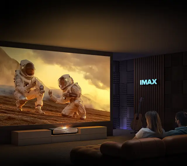 W ciemnym pokoju na dużym ekranie wyświetlany jest obraz dwóch astronautów na powierzchni planety. Na ścianie widoczne jest logo \'IMAX\'.